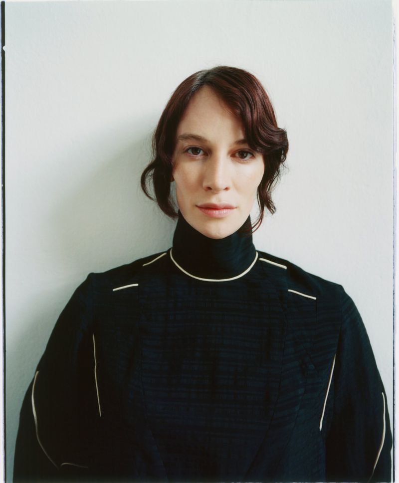 Portrait of an adult woman with auburn hair, wearing a black, high-necked top with white trim, against a plain white backdrop.