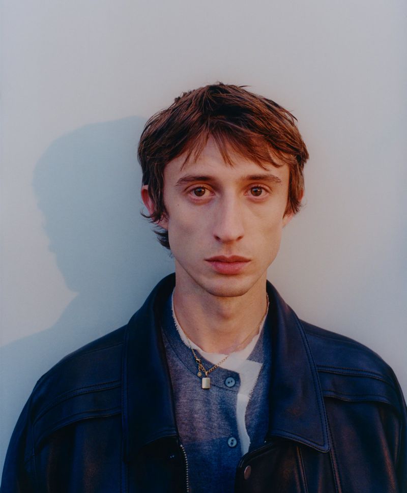 Headshot of a young adult male with fair skin and brown hair wearing a navy leather jacket, gray shirt, and gold necklace against a plain wall.