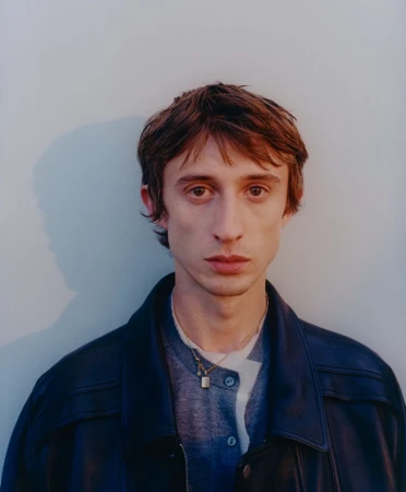 Headshot of a young adult male with fair skin and brown hair wearing a navy leather jacket, gray shirt, and gold necklace against a plain wall.