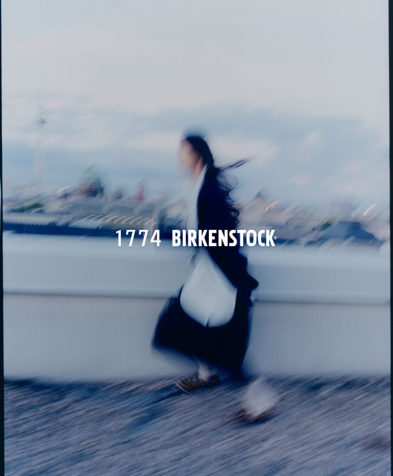 Fashion image of an adult woman in a black and white outfit walking outdoors against an urban backdrop.  Birkenstock brand visible.