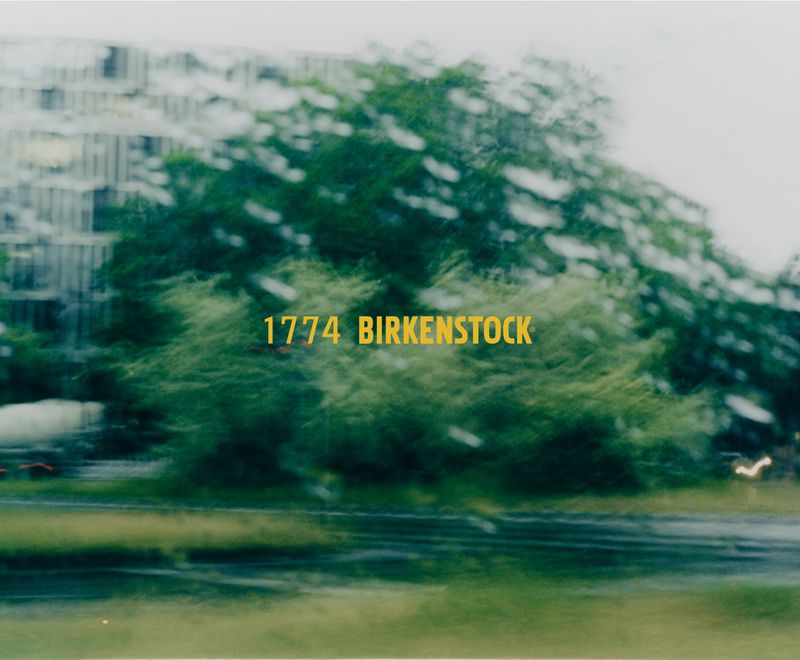 Abstract outdoor view through glass with motion blur showing blurred trees and yellow text '1774 BIRKENSTOCK' with building background.