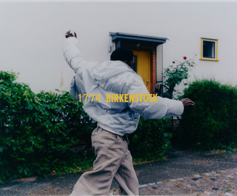 An adult throws a snowball in an urban setting with a house in the background. The person is wearing a white puffer jacket.