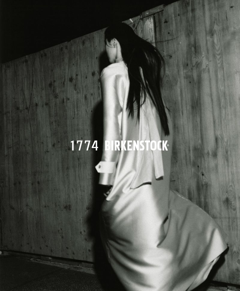 Monochrome fashion shot of a woman in a light-colored coat and skirt, standing in front of a weathered wooden wall with brand text overlay.