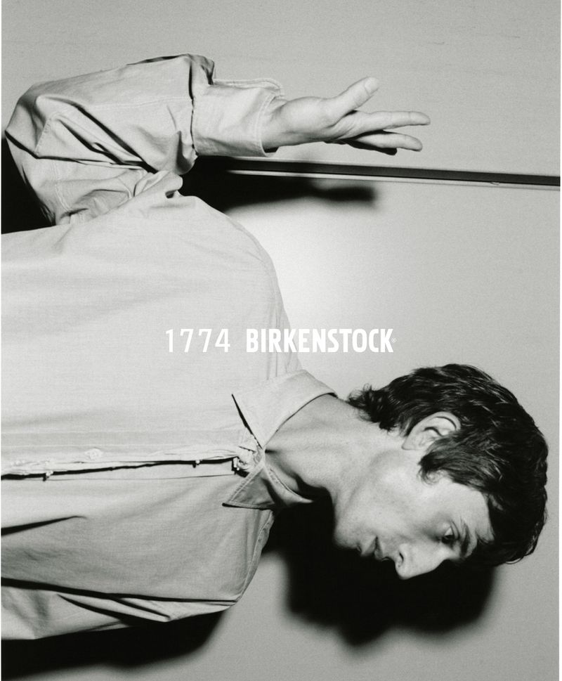 Monochromatic portrait of a man in a light-colored, long-sleeved shirt, facing right with raised hand and '1774 BIRKENSTOCK' text overhead.