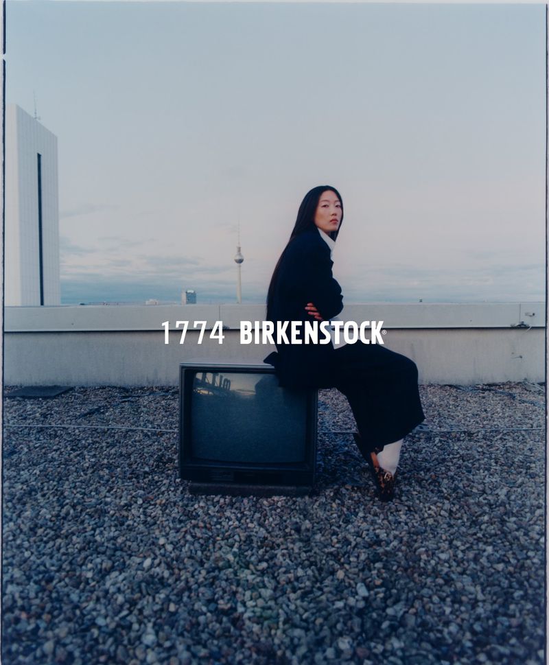 Asian woman in a dark business suit sitting on a vintage television on a rooftop with a city skyline in the background.