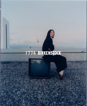 Asian woman in a dark business suit sitting on a vintage television on a rooftop with a city skyline in the background.