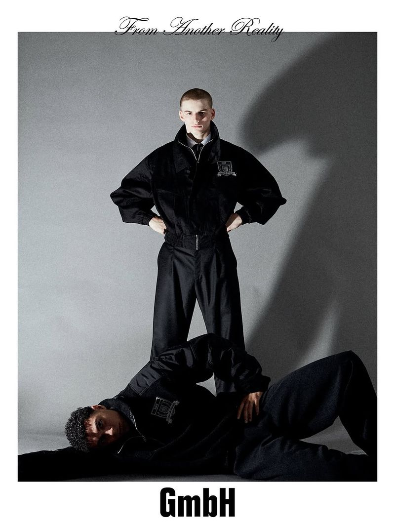 Two male fashion models pose in a studio. One model is standing, one is lying down. Both wear black bomber jackets and dark pants.