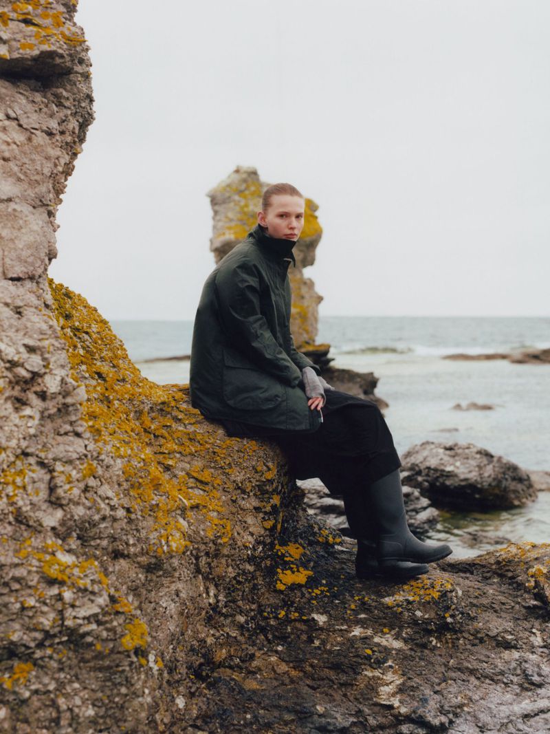 Adult wearing a dark green coat and black boots seated on the mossy rocks near the ocean with a large stone formation in the background.