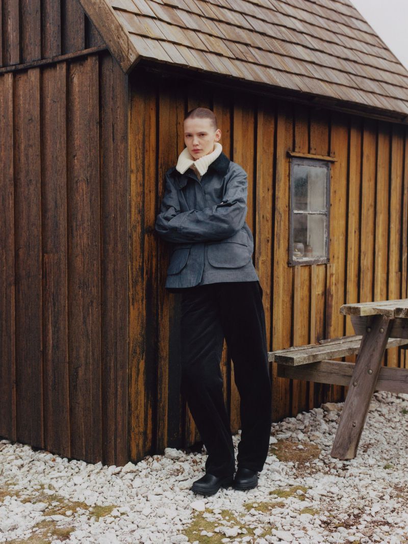 Adult male stands near a wooden shack in a grey jacket with fur collar, black pants and boots. Picnic table visible nearby.