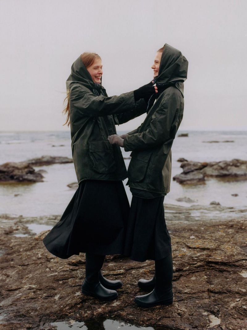 Two women in matching green hooded jackets and black skirts stand on a rocky beach. They are smiling, facing each other, against a cloudy sky.