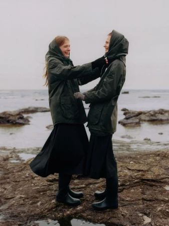 Two women in hooded olive green jackets and black skirts smiling together on a rocky coast