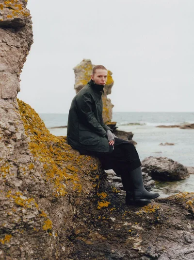 Model Agnes Lagercrantz sitting on a lichen-covered rock by the sea in a dark green wax jacket and black rain boots.
