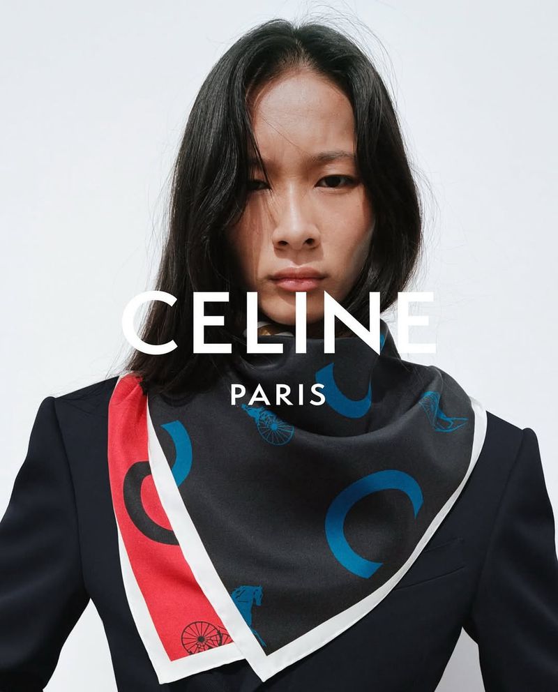 An Asian adult woman is wearing a black patterned silk scarf and a dark blazer, posed in front of a bright white seamless backdrop.
