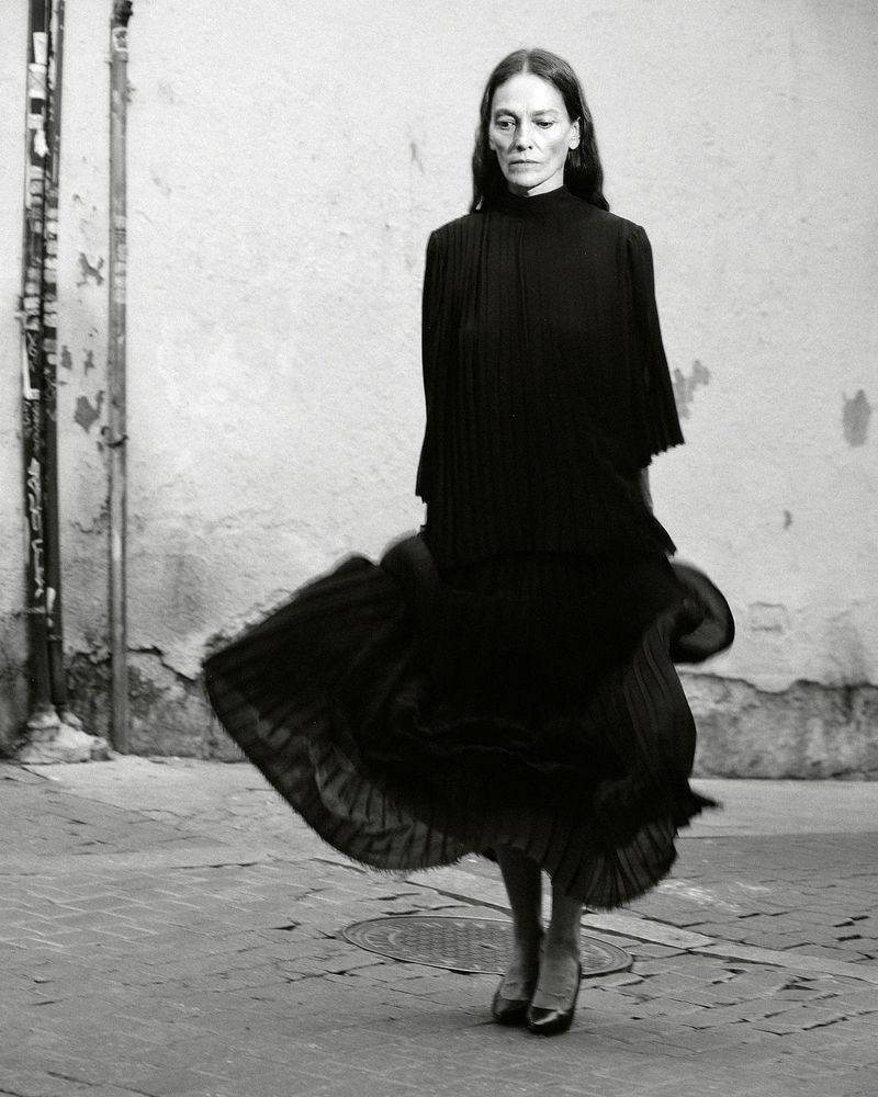 A woman in a dark dress stands in a plaza. She is wearing heeled shoes and has long hair. The photo is black and white with textured walls.