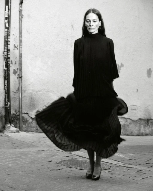 A woman in a dark dress stands in a plaza. She is wearing heeled shoes and has long hair. The photo is black and white with textured walls.