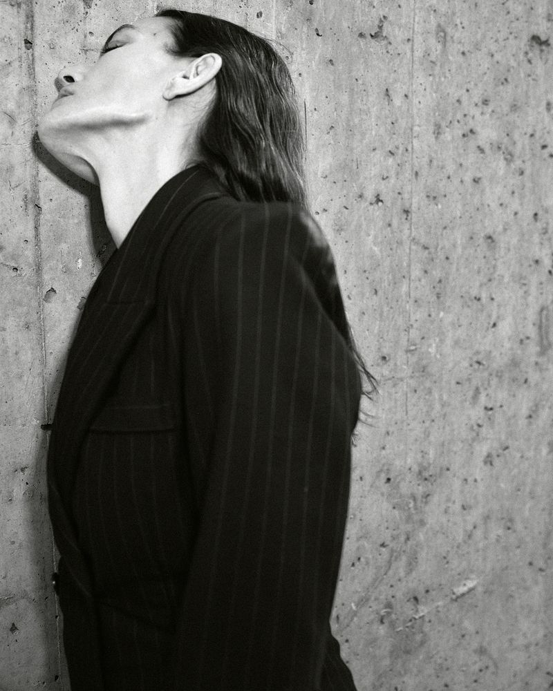 Monochrome portrait of a woman in a pinstripe blazer leaning against a textured concrete wall with eyes closed, wet hair and neutral expression.