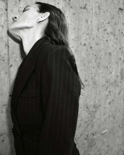 Monochrome portrait of a woman in a pinstripe blazer leaning against a textured concrete wall with eyes closed, wet hair and neutral expression.