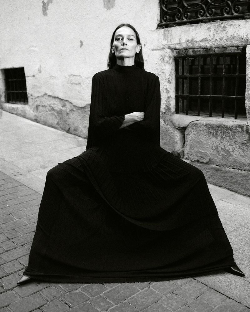 Elegant woman in a long black dress sits cross-legged in an outdoor urban alleyway with cobblestone and old stone walls in black and white.