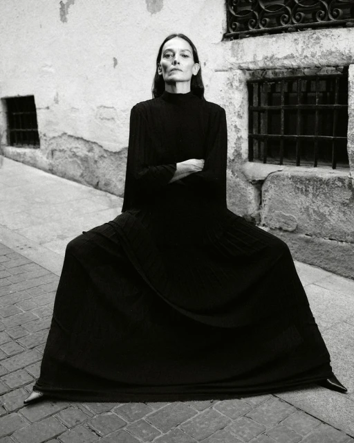 Elegant woman in a long black dress sits cross-legged in an outdoor urban alleyway with cobblestone and old stone walls in black and white.