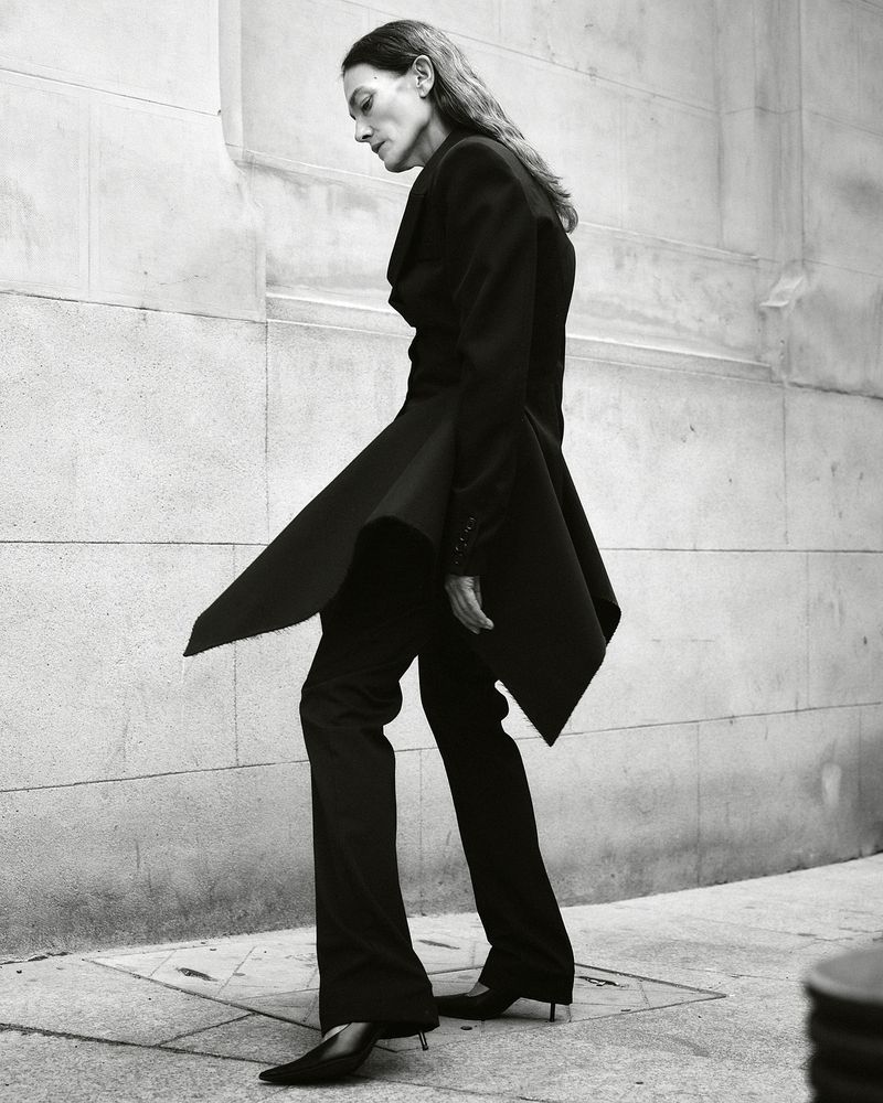 Model Laura Ponte in a black sculptural peplum suit and pointed heels walking outdoors against a stone building wall.