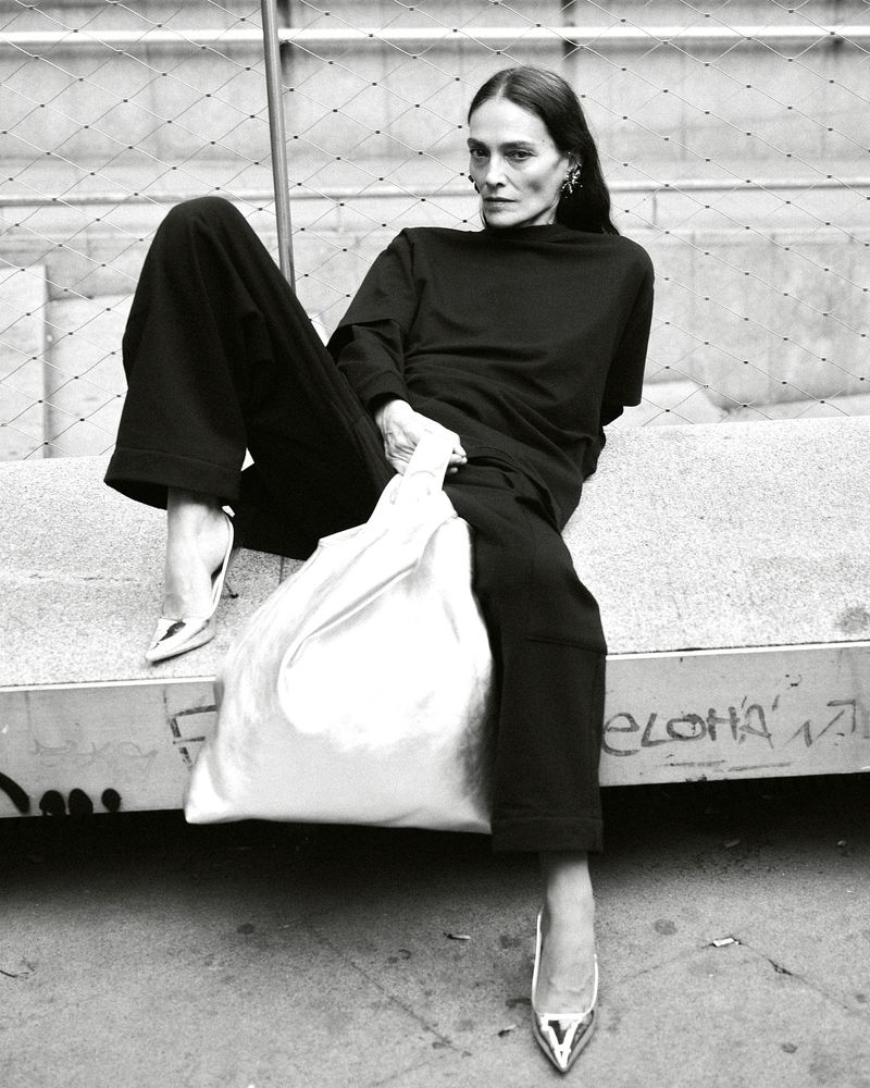 A tall, slim adult female model in a black outfit with metallic high heels poses while seated and holding a white shopping bag.