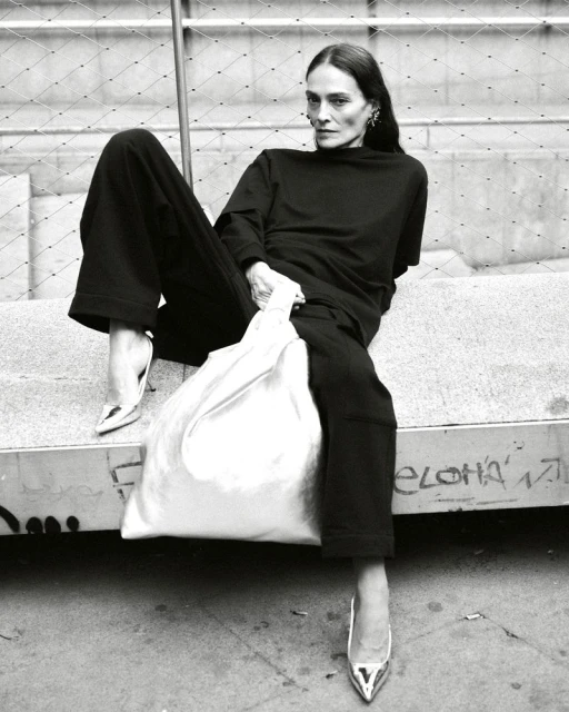 A tall, slim adult female model in a black outfit with metallic high heels poses while seated and holding a white shopping bag.