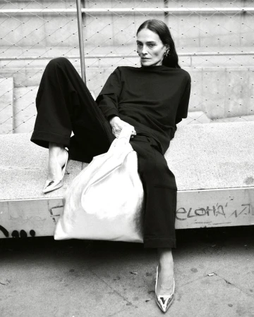 A tall, slim adult female model in a black outfit with metallic high heels poses while seated and holding a white shopping bag.