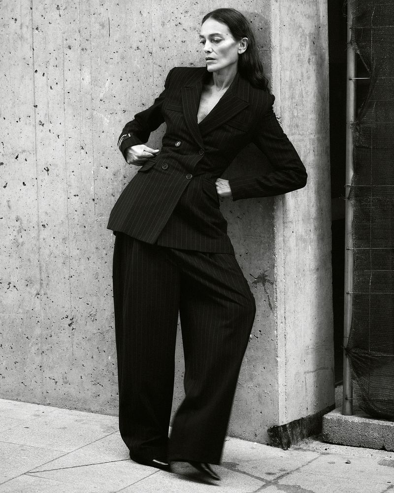 A black-and-white fashion shot of a woman in a pinstripe suit leaning against a concrete wall, posing with one hand on her blazer.