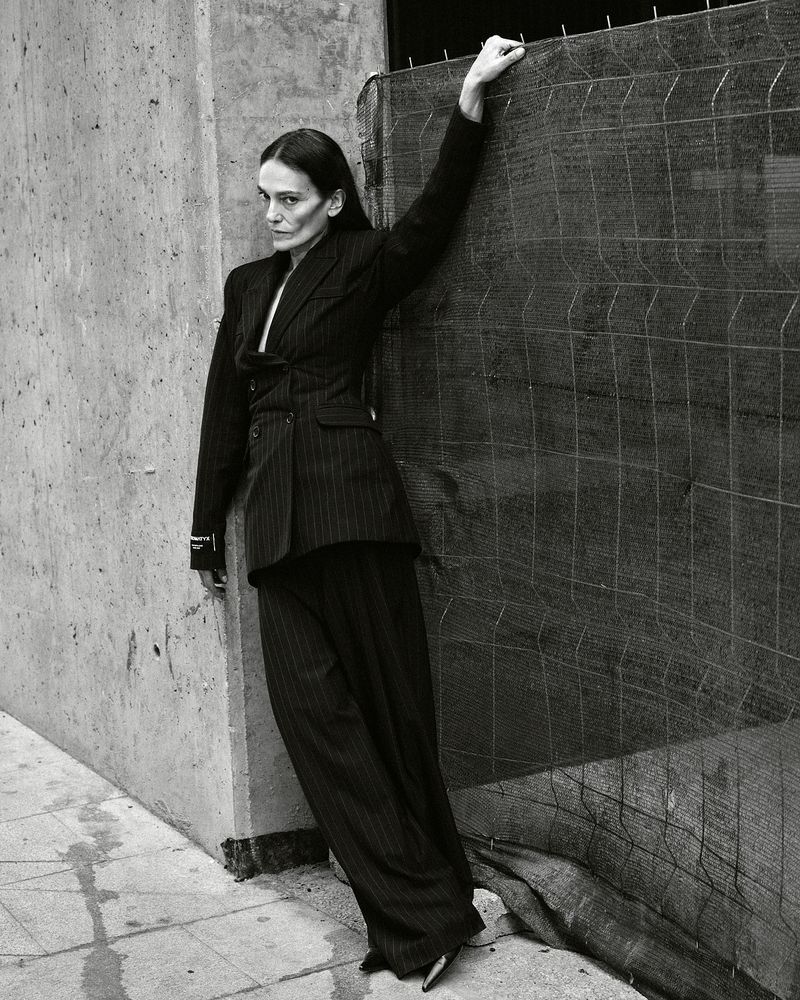 Elegant woman in a pinstripe suit poses confidently in an urban setting, leaning against a textured concrete wall and a wire mesh fence.
