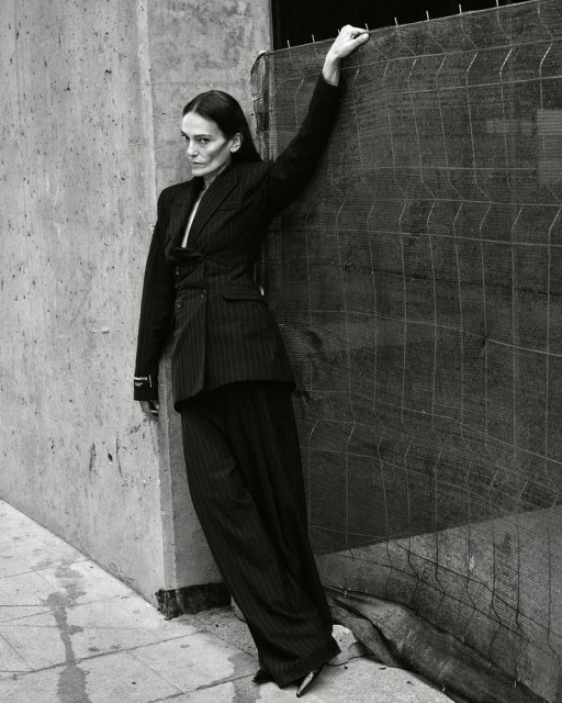 Elegant woman in a pinstripe suit poses confidently in an urban setting, leaning against a textured concrete wall and a wire mesh fence.