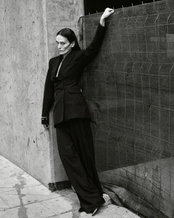 Elegant woman in a pinstripe suit poses confidently in an urban setting, leaning against a textured concrete wall and a wire mesh fence.