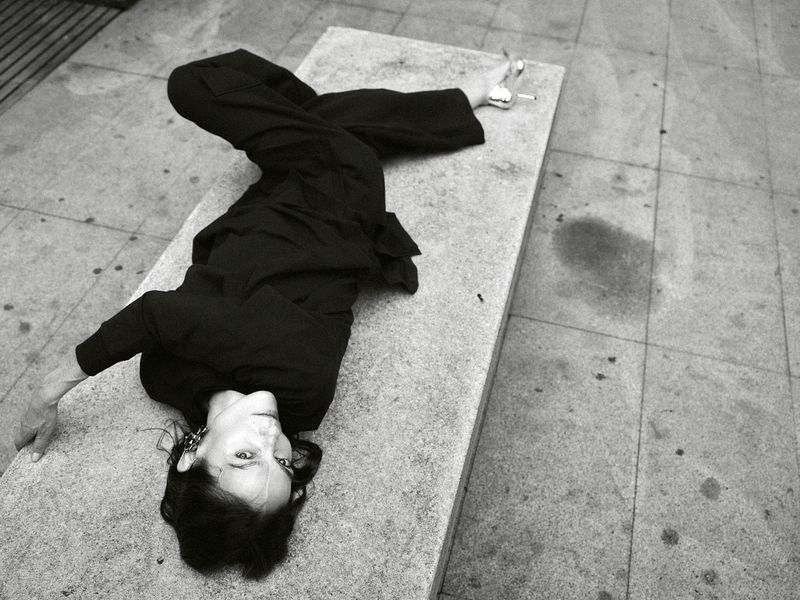 Woman in black outfit poses creatively on concrete bench outdoors in black and white, showing artistic expression and urban scene.