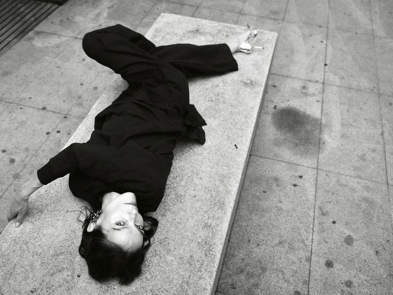 Woman in black outfit poses creatively on concrete bench outdoors in black and white, showing artistic expression and urban scene.