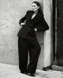 Model Laura Ponte leaning against a concrete wall wearing a dark pinstripe suit for Acromatyx Winter 25