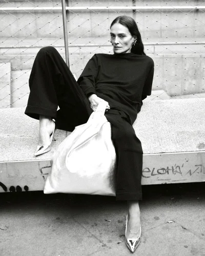 Model Laura Ponte seated outdoors holding a large metallic silver bag wearing black attire for Acromatyx Winter 25 by Javier Biosca.