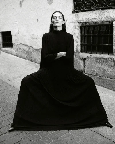 Model Laura Ponte seated on a cobblestone street wearing a black long-sleeved dress for ACROMATYX Winter 25