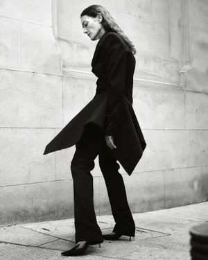Model Laura Ponte in a black sculptural peplum suit and pointed heels walking outdoors against a stone building wall.