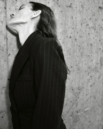 Black and white side profile of Laura Ponte in a dark pinstripe blazer leaning against a textured concrete wall.