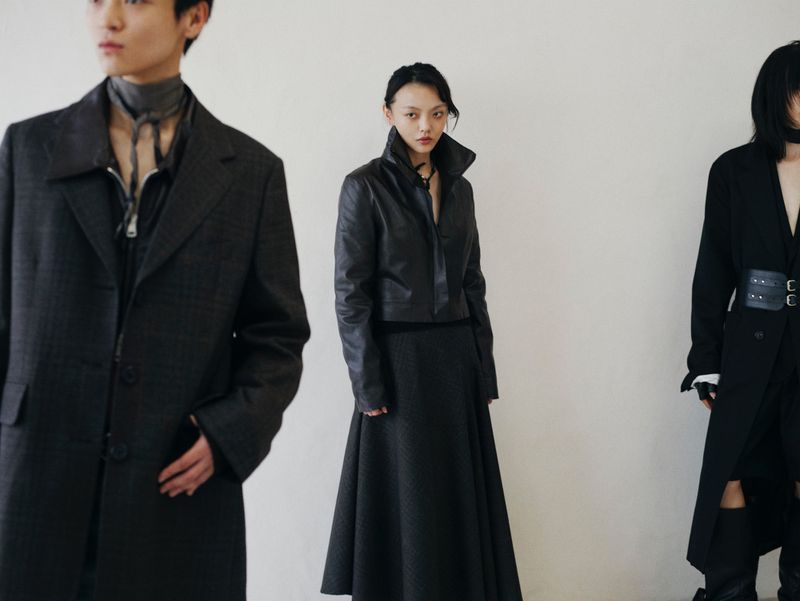 Three Asian fashion models pose in a photo studio wearing black coats, jackets, and dresses. The models look straight ahead. The background is plain white.