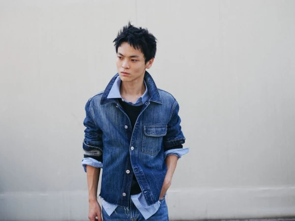 Model Yuki Koshiba Lin wears a layered blue denim jacket over a collared shirt against a plain wall for Our Legacy.