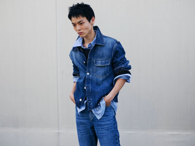 Young man in a denim jacket and blue jeans standing against a plain pale wall