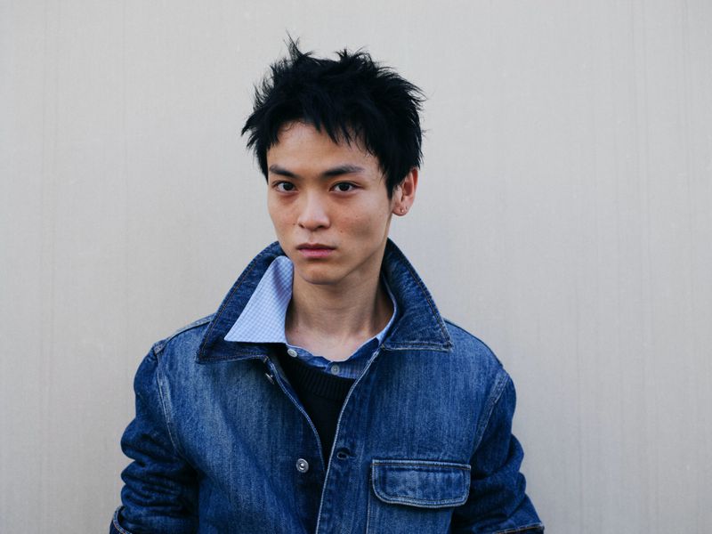 Close-up studio portrait of a young adult male with black hair, wearing a denim jacket and buttoned shirt against a plain wall.
