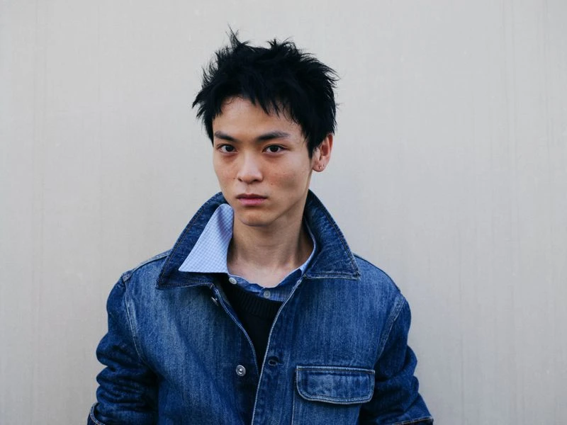 Close-up studio portrait of a young adult male with black hair, wearing a denim jacket and buttoned shirt against a plain wall.