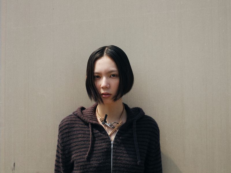 Portrait of an East Asian adult woman with bob haircut wearing hooded sweater and statement necklace against a beige wall.