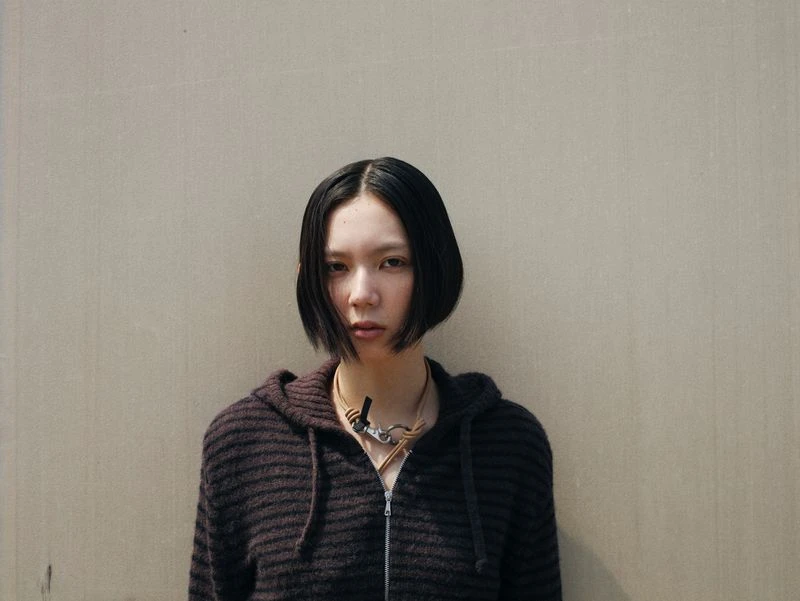 Portrait of an East Asian adult woman with bob haircut wearing hooded sweater and statement necklace against a beige wall.