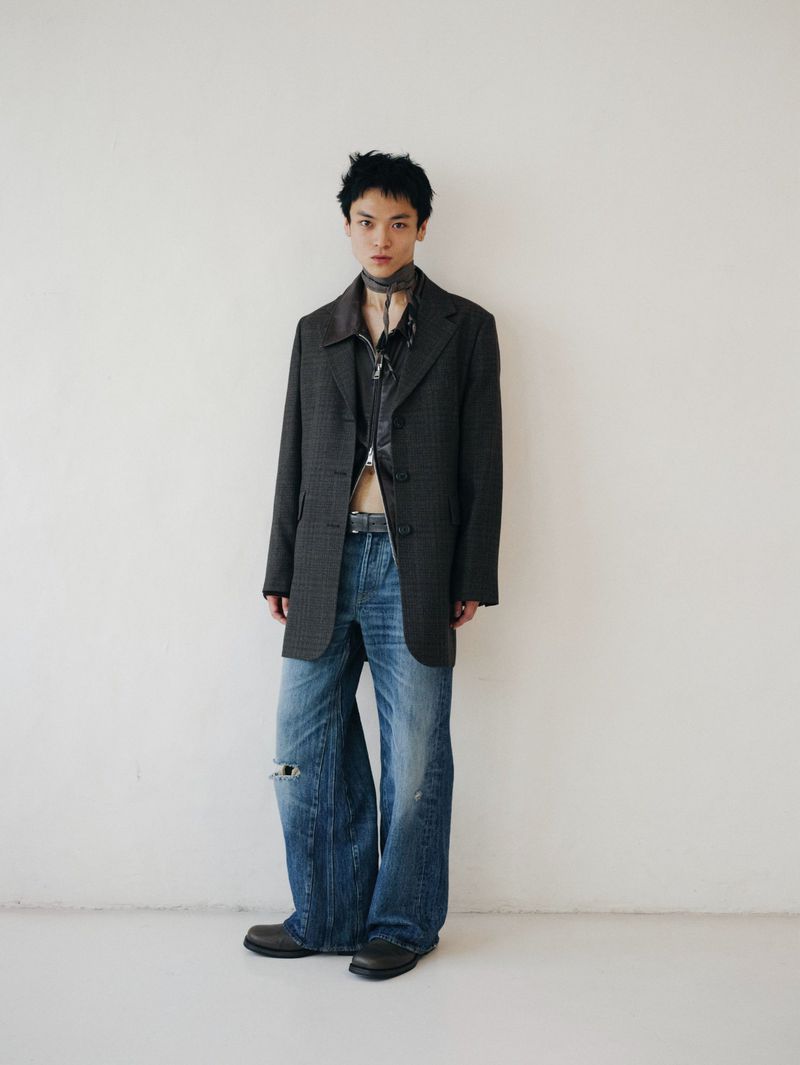 A young adult male stands against a white wall wearing an oversized grey blazer, black top, ripped blue jeans, and black shoes.