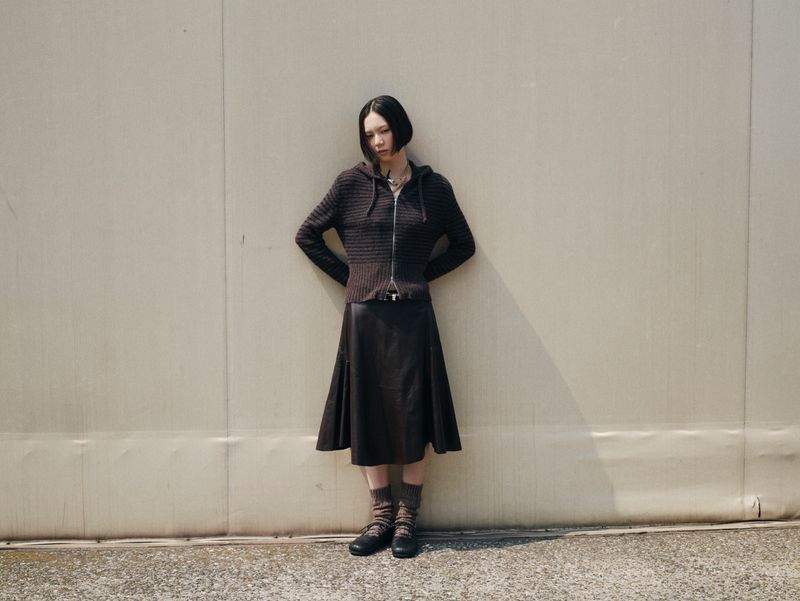Adult woman poses against a grey wall in a dark sweater, skirt, socks, and shoes. The natural light creates shadows on the background.