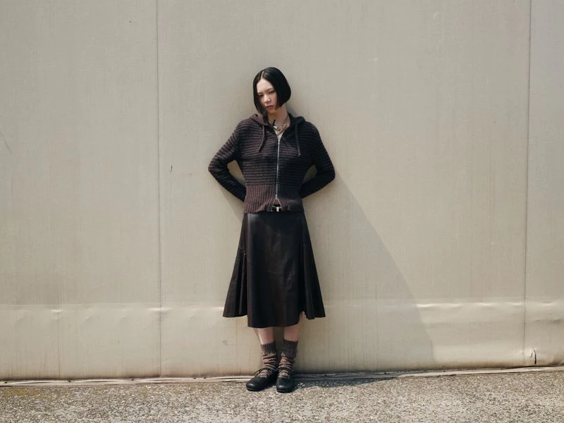 Adult woman poses against a grey wall in a dark sweater, skirt, socks, and shoes. The natural light creates shadows on the background.
