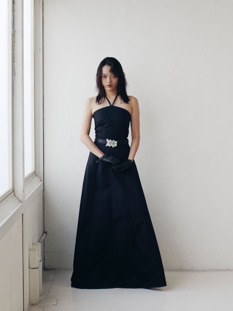 Woman in a black halter dress and black gloves stands near a bright window. Fashion shot in a minimalist white room.