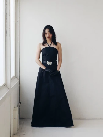 Woman in a black halter dress and black gloves stands near a bright window. Fashion shot in a minimalist white room.